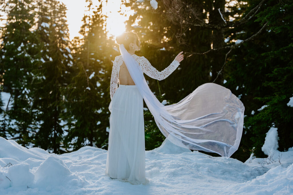 Bride wearing backless elopement dress with veil. It's sunset, she's surrounded by trees and snow because it's winter, and she's on Grouse Mountain in Vancouver