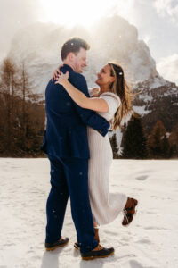 Passo Gardena Dolomites Winter Engagement, couple smiling at each other as the snow falls with the sun over Sassolungo