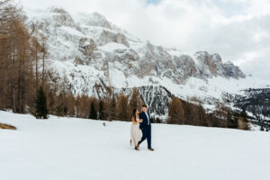 Passo Gardena Dolomites Winter Engagement, couple walks through the snow with the Odle mountains behind them