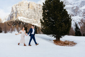 Passo Gardena Dolomites Winter Engagement, man and woman skipping passed tree in the snow