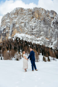 Passo Gardena Dolomites Winter Engagement, couple looking back at camera while walking towards mountains