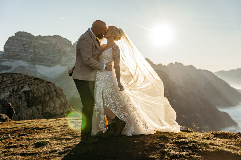Bride wearing thin strapped summer dress with long veil and groom wearing beige suit on mountain top of Cadini di Misurina