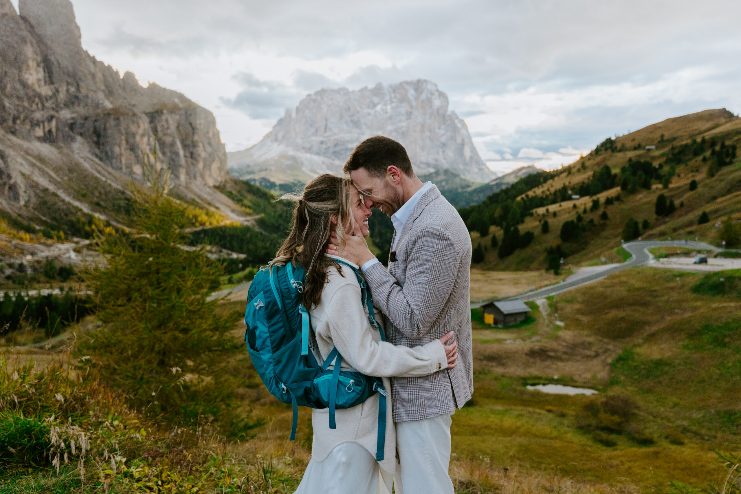A couple shares an intimate moment during their elopement at Passo Gardena in the Dolomites. The groom, in a light-patterned blazer, holds the bride's face as they touch foreheads. The bride wears a white dress and a teal hiking backpack, standing against a backdrop of dramatic limestone peaks and winding mountain roads.