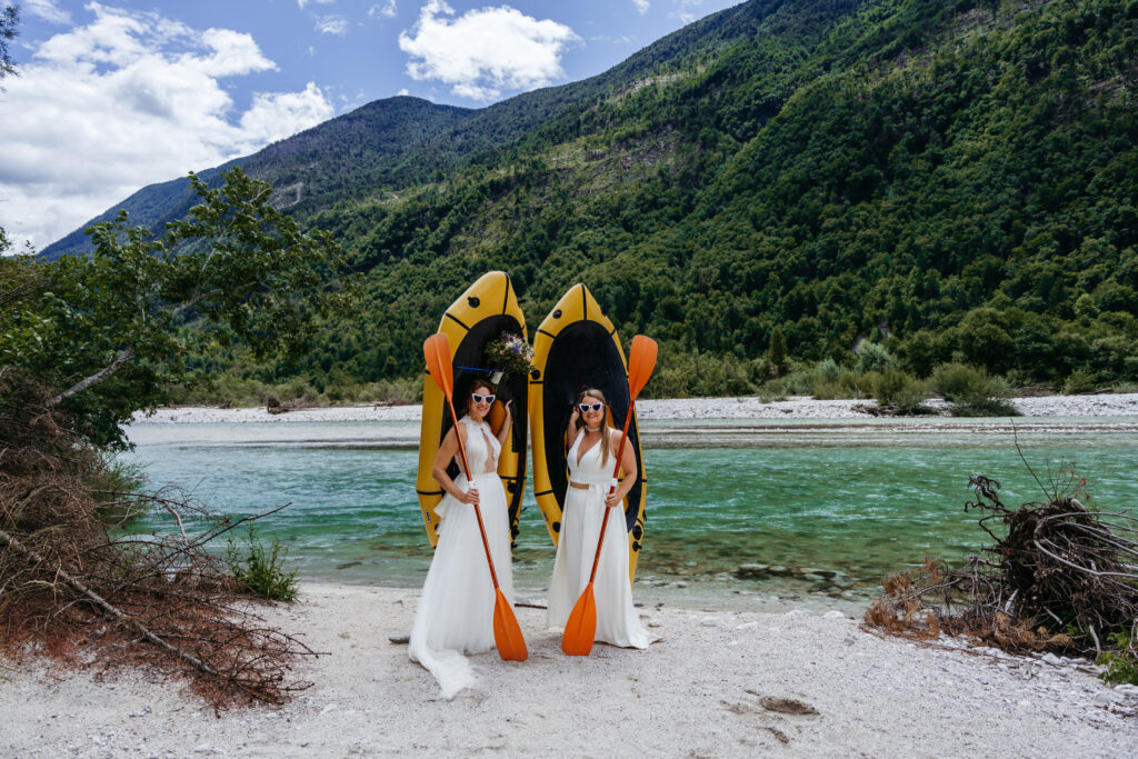 Two brides in white dresses and sunglasses hold paddles in front of yellow kayaks on a rocky riverbank, with green mountains in the background.