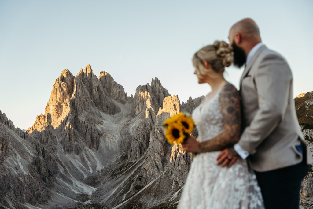 A couple stands together in soft focus during a mountain elopement, overlooking dramatic rocky peaks at golden hour. The bride wears a lace wedding dress and holds a bouquet of sunflowers, while the groom wears a light beige suit. Warm sunlight highlights the rugged alpine landscape in the background.