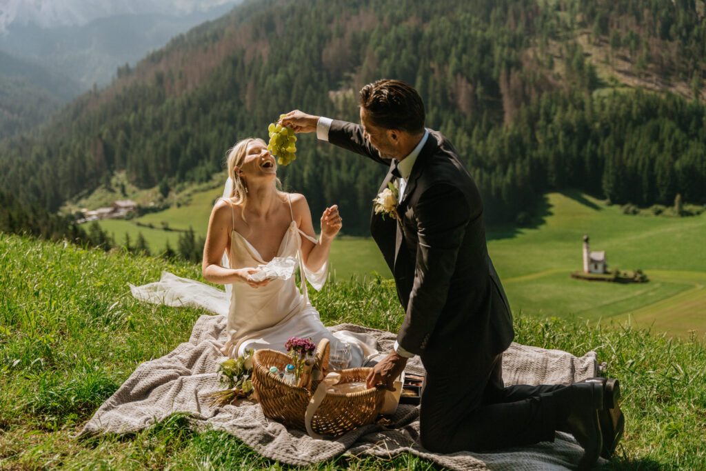 Couple enjoying a picnic on a grassy hillside in Val di Funes, Italy, overlooking a sunlit mountain valley. The groom playfully holds a bunch of green grapes above the laughing bride while they sit on a blanket beside a wicker picnic basket, with the small Chapel of St. Johann and forested hills in the background.