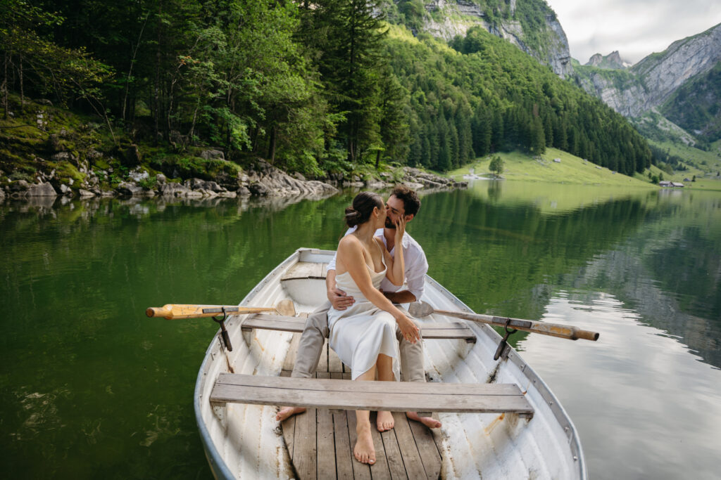 Couple embracing and kissing in a small rowboat on a calm emerald lake surrounded by forested mountains in Switzerland. The bride wears a simple white dress while the couple sits barefoot in the center of the boat, with soft reflections of the trees and cliffs mirrored in the still water.