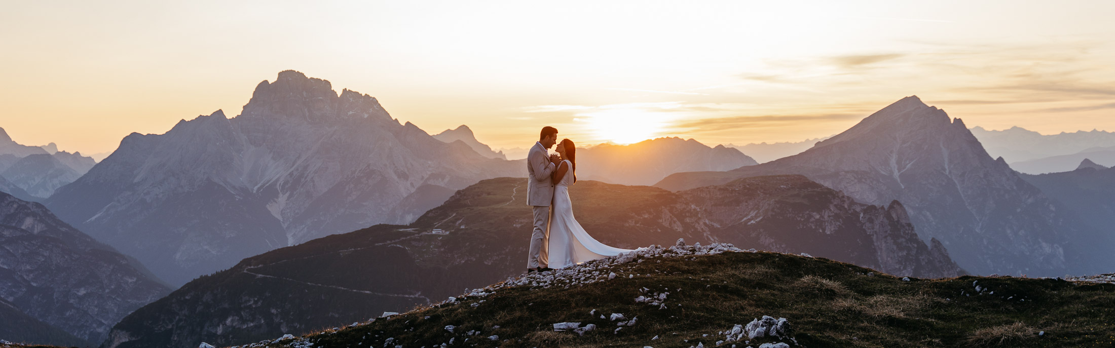 Bride and groom standing holding hands and facing each other with the Dolomites and setting sun in the background