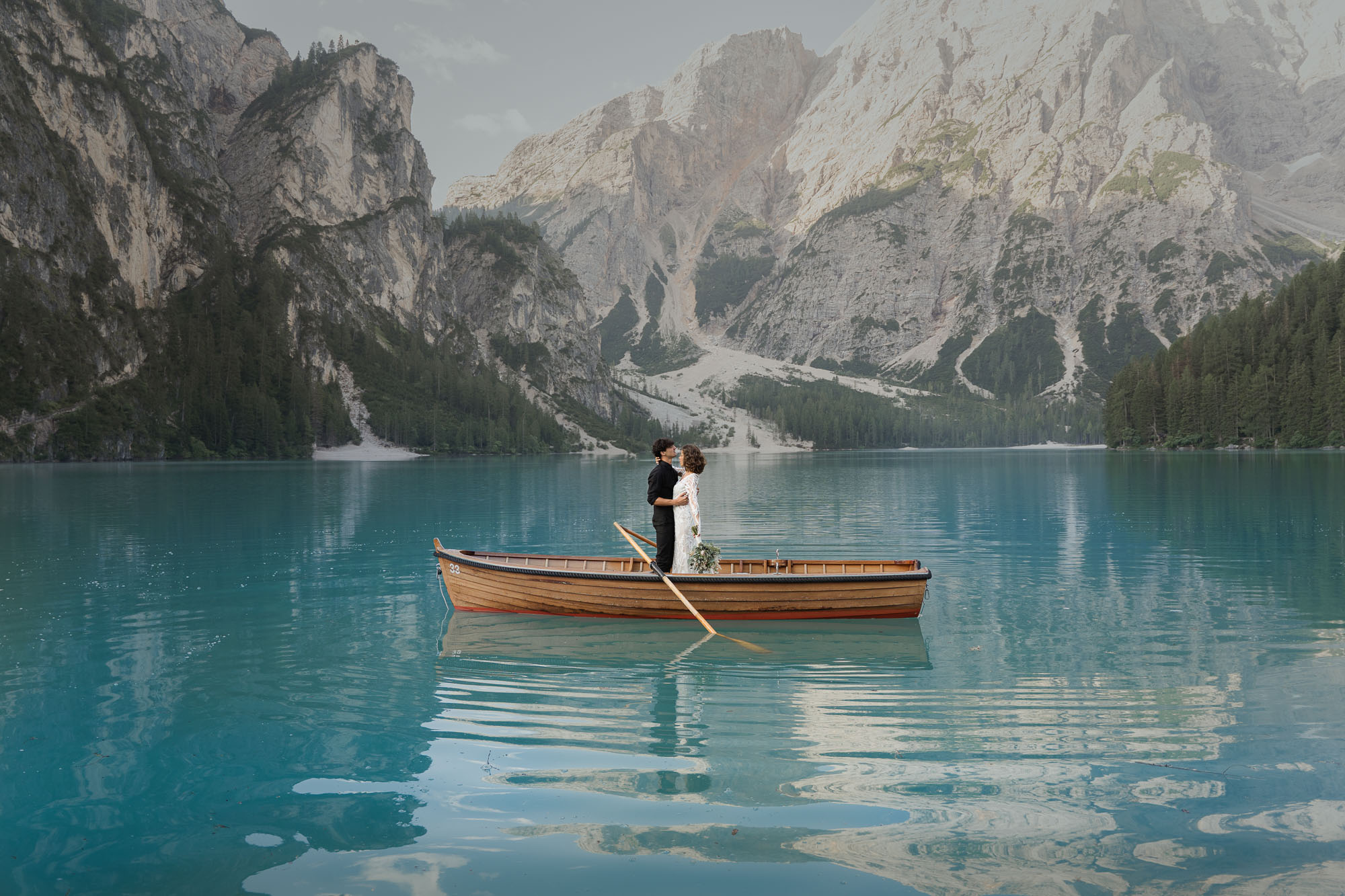 Bride and groom standing on rowboat on Lago di Braies rented from La Palifitta with mountain in the background at sunrise