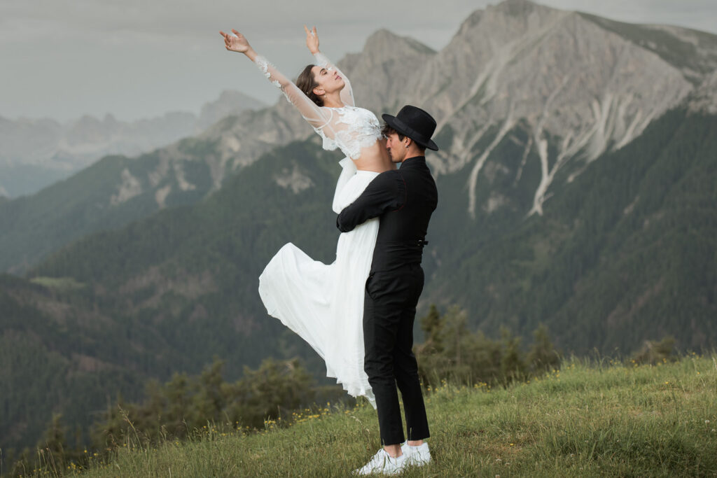 A couple stands on a grassy mountain overlook during an adventure elopement. One partner lifts the other into the air as they extend their arms and smile, with rugged mountain peaks and evergreen forests stretching into the background.