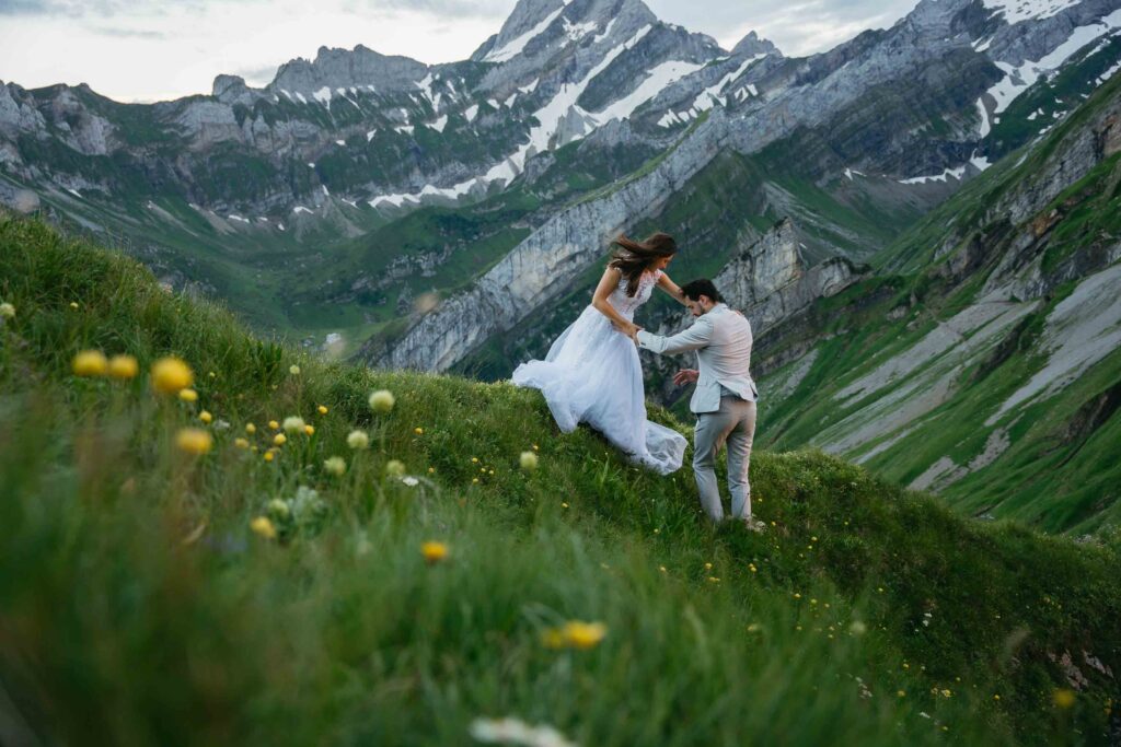 A couple in wedding attire walks hand in hand along a grassy alpine ridge during an adventure elopement. One partner helps the other up the hillside as dramatic, snow-dusted mountain peaks and wildflowers surround them.