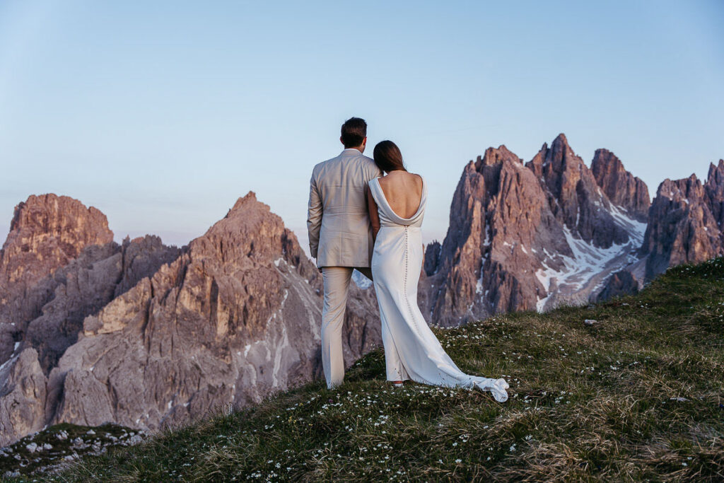 A newlywed couple stands arm in arm on a grassy mountain ridge, facing a dramatic range of jagged peaks at sunset. The bride’s long white dress trails behind her as they take in the sweeping alpine landscape together during their adventure elopement.