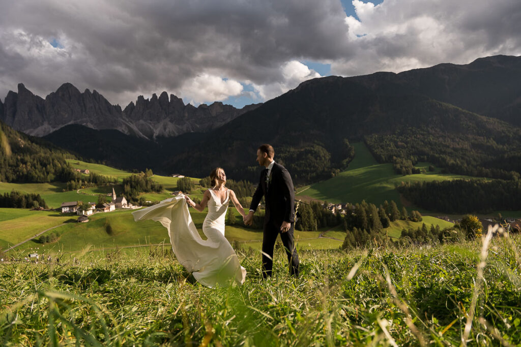 Wife and husband frolicking through fields of Val di Funes in the Dolomites after their vow renewal