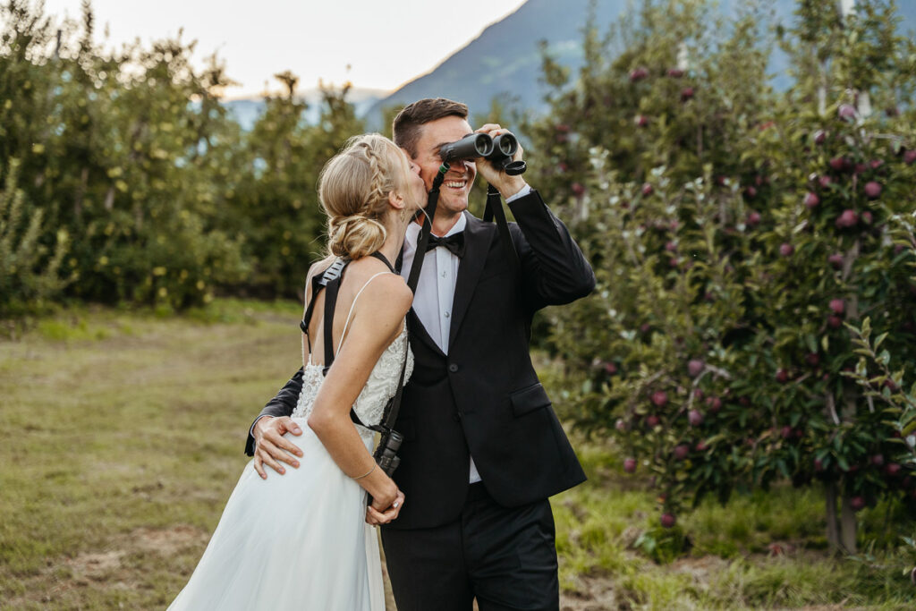 A newlywed couple stands in an apple orchard during their elopement day. One partner looks through binoculars while the other leans in smiling, with mountains and rows of apple trees in the background.