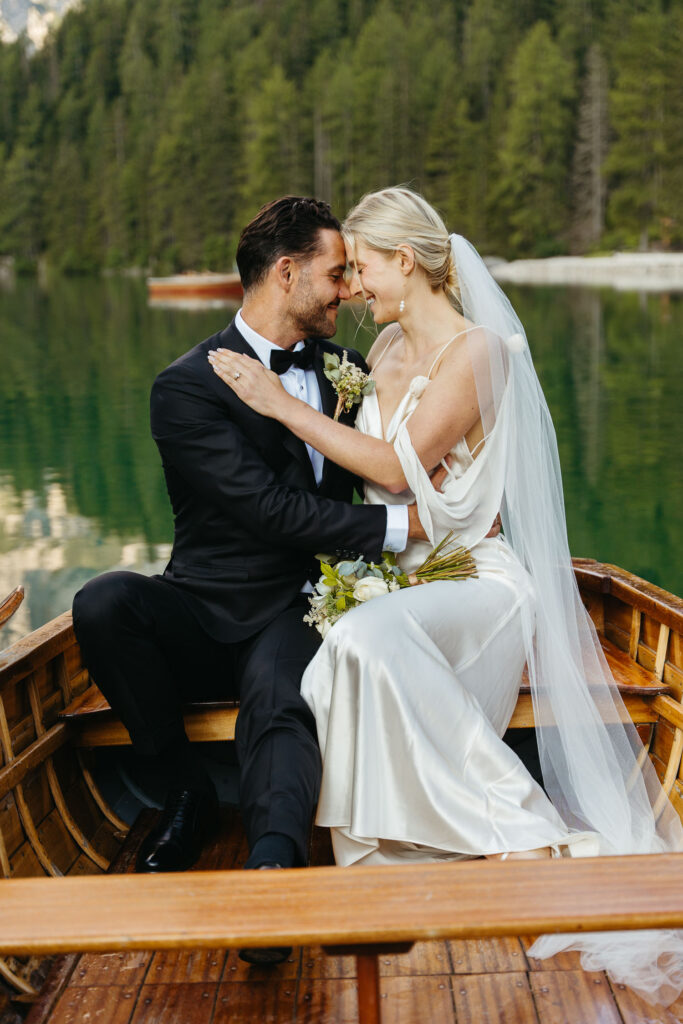 Bride and groom in a rowboat at Lago di Braies with trees in the background. Bride is wearing a lightweight elopement dress from silk or satin and groom is wearing a black suit with bowtie