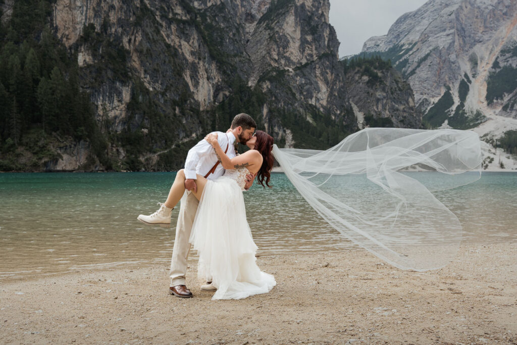 Groom holding bride in a leaned-back dancer position with her veil flowing in the air, and Lake Braies in the background. Bride is wearing a strapless elopement dress, groom is wearing a white shirt with suspendors
