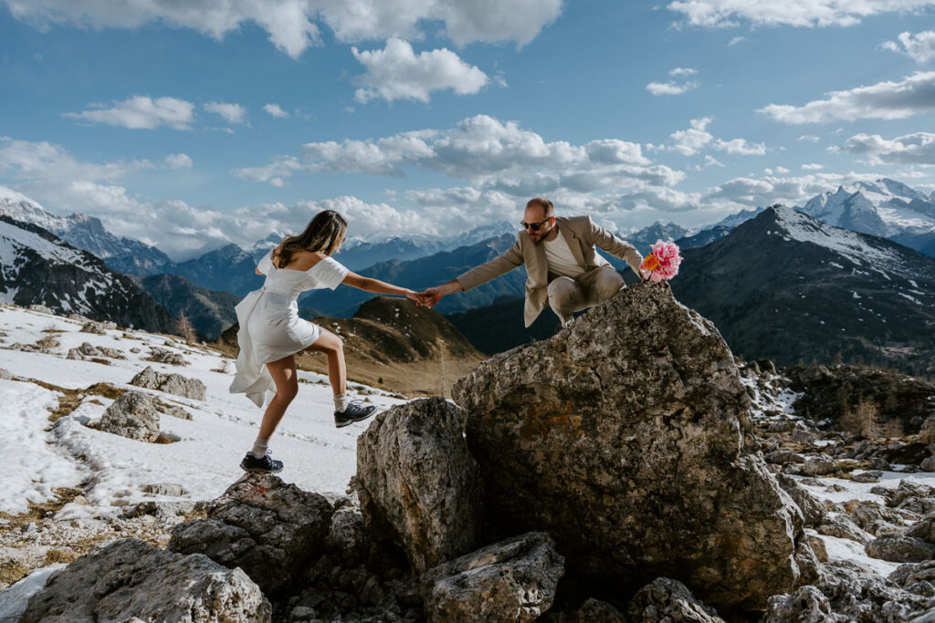 Bride wearing short elopement dress with long extention, groom wearing beige suit and helping bride stand on a rock. They are in the snow at Passo Giau in spring in the Dolomites