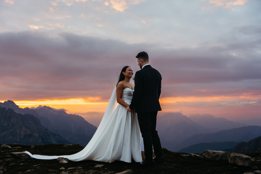 Adventure elopement of newly eloped couple in wedding attire standing together on the rocky mountain peaks of Tre Cime at sunset, surrounded by dramatic alpine scenery and soft golden light.