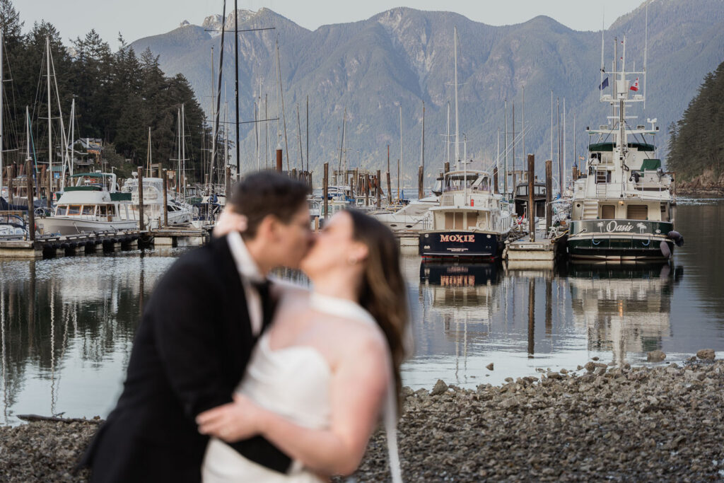 Bride and groom kissing out of focus with Bowen Island boats and mountains in the background of their cool summer elopement as they avoid the heatwaves and fires