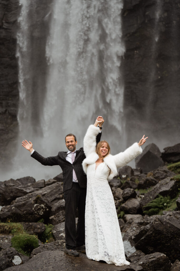 Bride and groom super happy about getting married with hands in the air and Fossà behind them on Faroe Islands