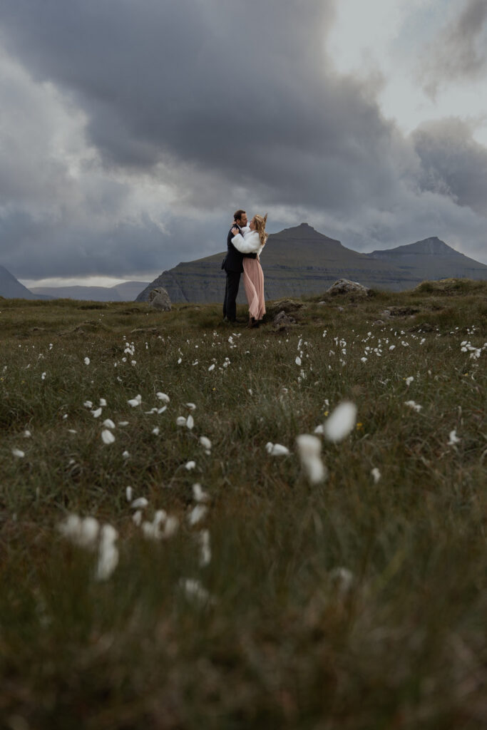 Bride and groom learning how to elope on the Faroe Islands with fluffy plants in the forground and dramatic cliffs in the background.