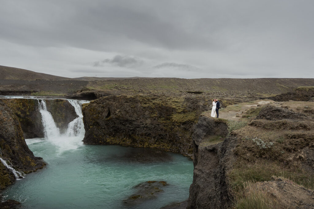 Bride and groom standing on a cliff above Sigoldufoss. They wanted to book a cool summer elopement destination to avoid heatwaves and wildfires, so they chose to elope in Iceland