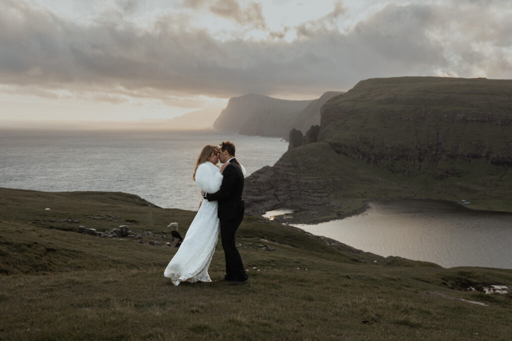 Bride and groom embracing on Faroe Islands with sunset in the background at Traelanipa. Ocean and lake in view and seacliffs. How to elope in the Faroe Islands: Ultimate Guide.