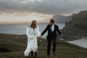 Bride and groom running towards the camera as the sun sets behind the mountain leaving a warm glow at Trælanípa. This is their Faroe Island elopement. Sheep in the backgound