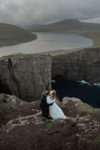 Bride and groom with steep cliffs below them overlooking the lake of Trælanípa and the ocean on Faroe Islands, for their elopement and vow renewal.