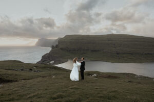 Bride helping groom get ready for their elopement on Faroe Isalnds at Trælanípa, with the lake and ocean and sunset in the background