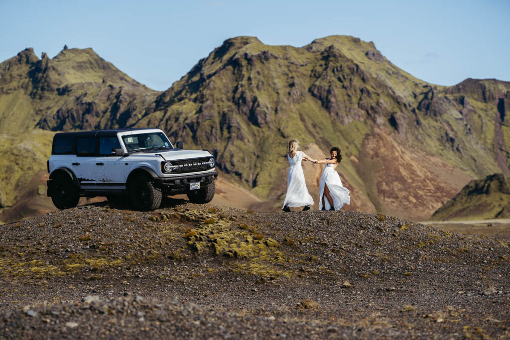 Two brides spinning in circles with their 4x4 and Icelandic hills in the background; How to Elope in Iceland