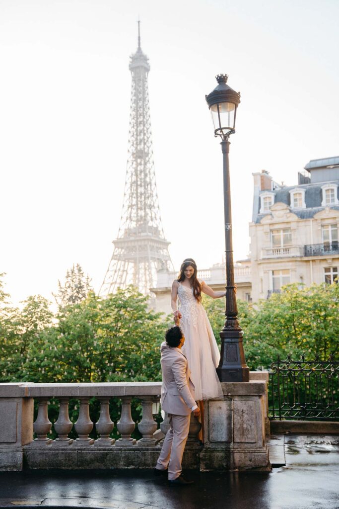 Bride and groom at sunrise with the Eiffel Tower in the background, they are looking at each other and she is standing on a brick wall while holding his hand; How to Elope in Paris 