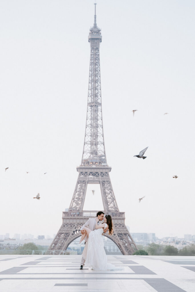 Bride and groom dancing and kissing at the Eiffel Tower at sunrise with pigeons flying around; How to Elope in Paris
